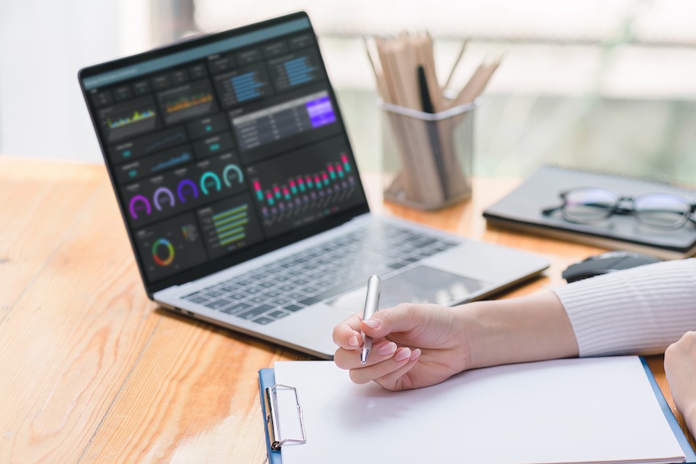 Cropped Shot Of A Professional Female Doctor Is Taking Notes In A Medical Journal And Filling Out Documents, Including The Patient's Illness History, Looking At A Laptop Screen Dashboard.