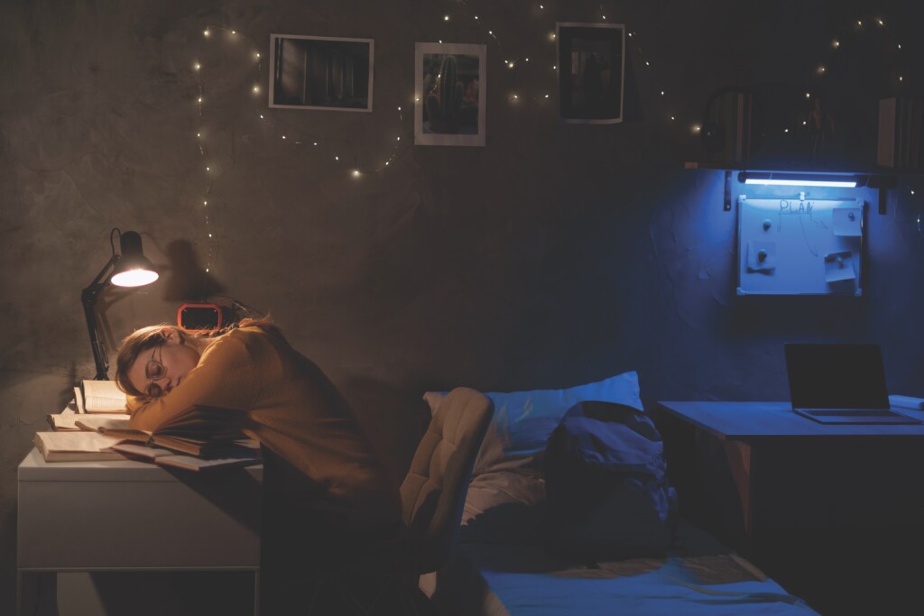 Tired College Student Sleeping At Table In Dormitory Preparing For Exam At Late Night.
